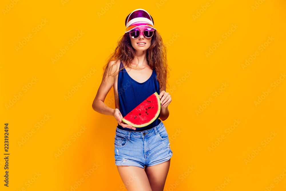 Beautiful young female in cap, sunglasses and swimsuit hold slice of watermelon and looking away while standing on yellow background. Copyspase