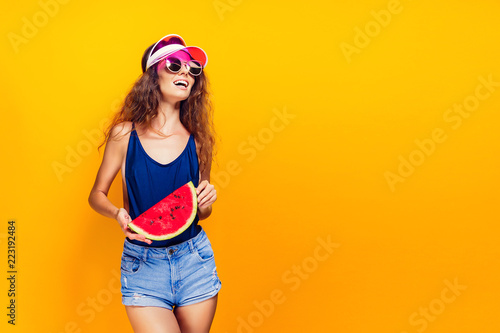 Beautiful young female in cap, sunglasses and swimsuit hold slice of watermelon and looking away while standing on yellow background. Copyspase
