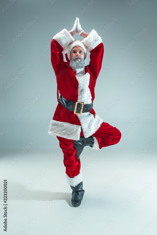 Funny serious guy with christmas hat sitting at yoga pose at studio. New Year Holiday. Christmas, x-mas, winter, gifts concept. Man wearing Santa Claus costume on gray. Copy space. Winter sales.