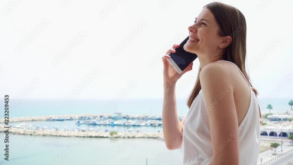 Smiling young woman in white dress using mobile on balcony. Female on vacation enjoying sea view from terrace in morning and talking to friend via cellphone in slow motion Smiling young woman in white dress using mobile on balcony. Female on vacation enjoying sea view from terrace in morning and talking to friend via cellphone in slow motion
