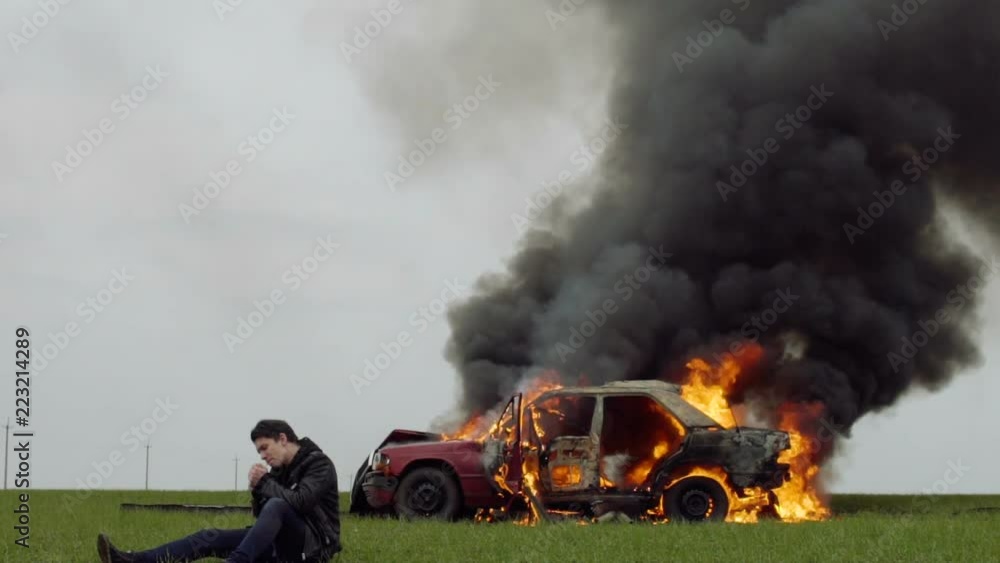 A guy lights a cigarette against the backdrop of his burning car, the harm of smoking, a man smokes against the background of a fire car, slow-mo A guy lights a cigarette against the backdrop of his burning car, the harm of smoking, a man smokes against the background of a fire car, slow-mo