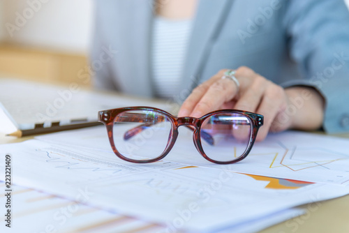 closeup eyeglasses and female hand on background laptop and graph report data on wood desk working at the office