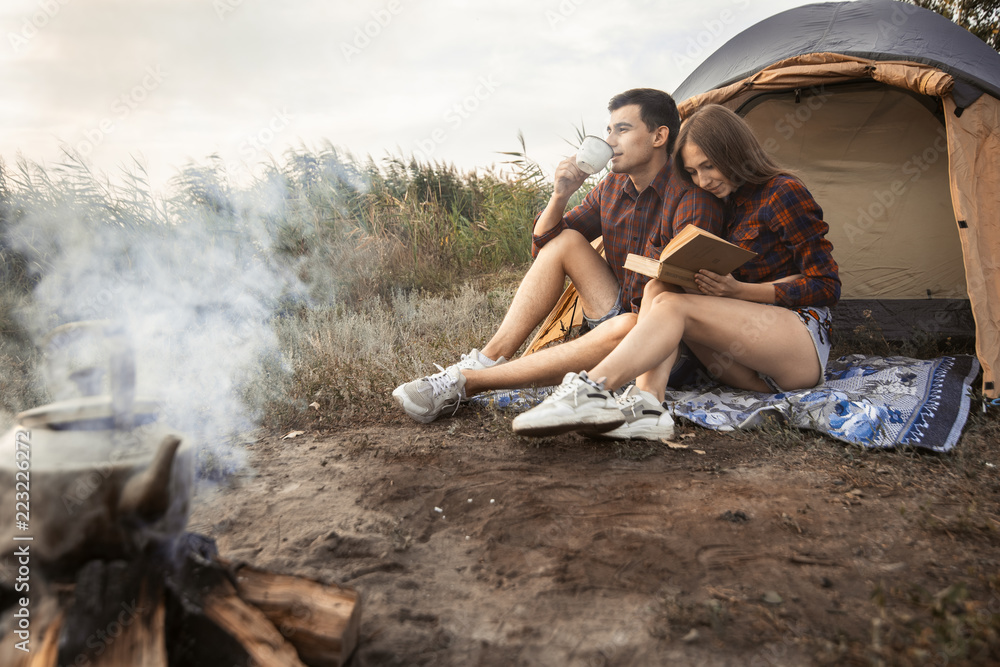 beautiful guy and girl dressed in red checkered shirts sitting on the ground near a tent and a fire