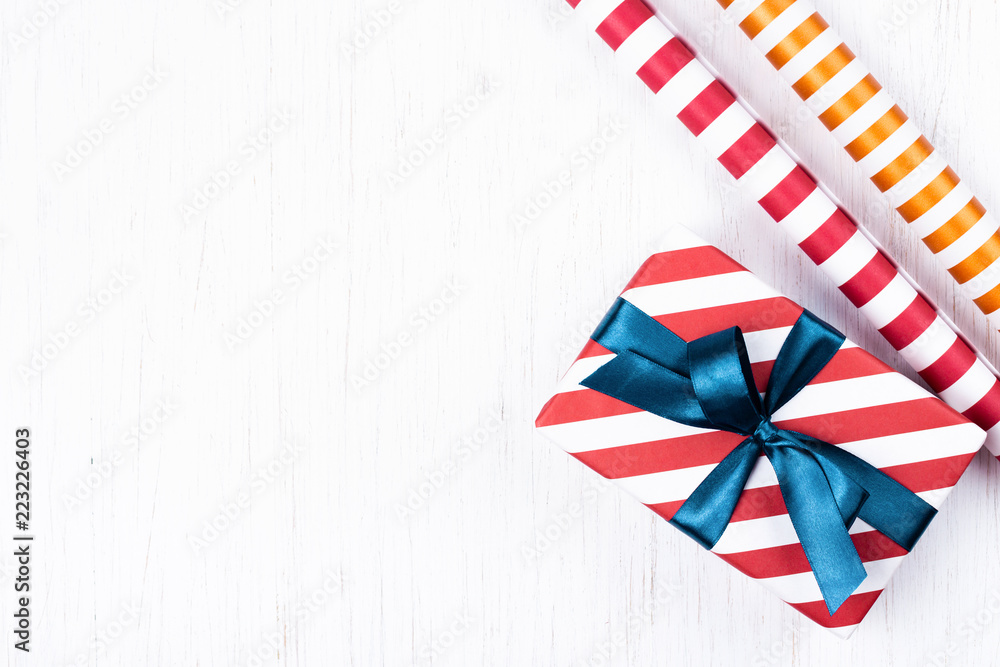 Christmas gift wrapped in striped gift paper tied with blue ribbon and two decorative paper rolls on white wooden background. New Year, holidays and celebration concept, top view with copy space. Christmas gift wrapped in striped gift paper tied with blue ribbon and two decorative paper rolls on white wooden background. New Year, holidays and celebration concept, top view with copy space.