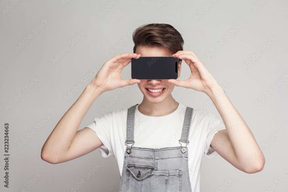 Happy modern young boy in demin overalls and white T-shirt standing, holding and covering eyes with smartphone, like eyeglasses and toothy smile. Indoor, isolated, studio shot, grey background Happy modern young boy in demin overalls and white T-shirt standing, holding and covering eyes with smartphone, like eyeglasses and toothy smile. Indoor, isolated, studio shot, grey background