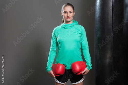 Portrait of a confident young athlete woman with collected hair standing near punching bag, holding boxing red gloves and looking at camera. Indoor studio shot, isolated on dark grey background.