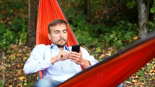 Young handsome man using smartphome while sitting in hammock in autumn forest. photo