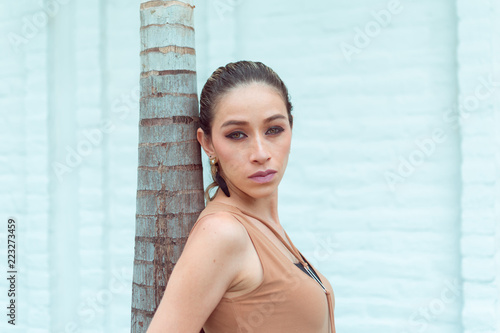 a beautiful slim woman resting on a palm tree staring at the camera, has green eyes and good makeup, photographed in the foreground, model located on the side of the composition