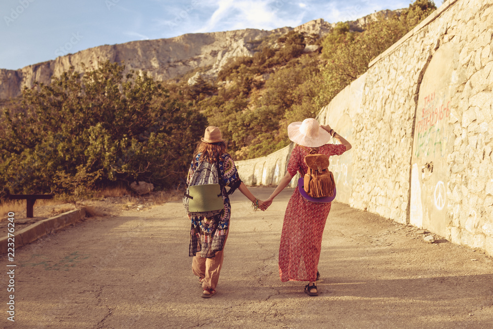 Fototapeta premium Girlfriends-travelers in the style of a jeeps are holding hands, brightly dressed with bonnies on their heads, walking along the road. In the background, mountains and forest. The concept of freedom.
