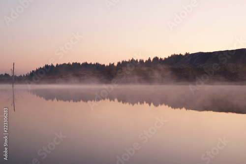 Landscape early morning on the lake with fog and reflection of forest and hills on a surface of water.
