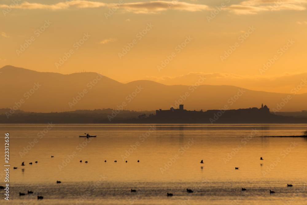 Beautiful view of Trasimeno lake (Umbria, Italy) at sunset, with orange tones, birds on water, a man on a canoe and Castiglione del Lgo town on the background