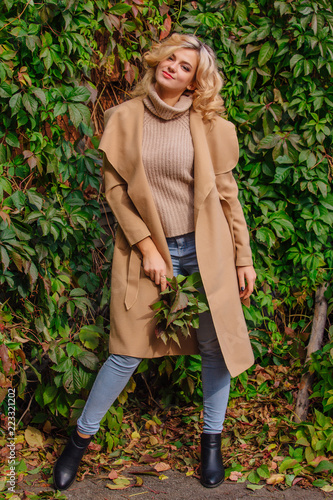 Young beautiful woman in sweater and coat stands next to the background of wild grapes holding bouquet of autumn leaves