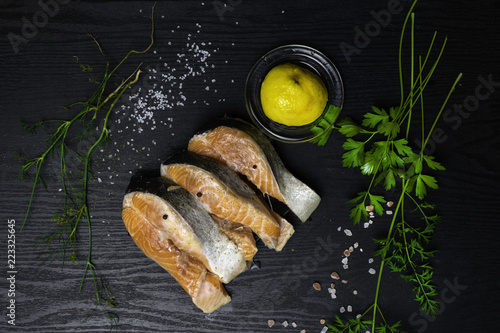 Slices of red fish salmon on trout with lemon, parsley, dill, pink and white salt, selery greens on black wooden background with copy space for your text. Top view photo