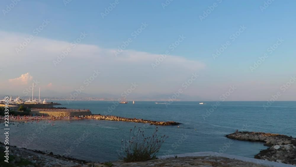 Harbour (Port Vauban) and beach (Plage de la Gravette) in Antibes, southern France. Stone wall, sea, horizon and cloudy sky showing. Small plant in foreground. Establishing shot. Harbour (Port Vauban) and beach (Plage de la Gravette) in Antibes, southern France. Stone wall, sea, horizon and cloudy sky showing. Small plant in foreground. Establishing shot.
