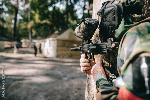 selective focus of paintball player in goggle mask and camouflage aiming by paintball gun outdoors