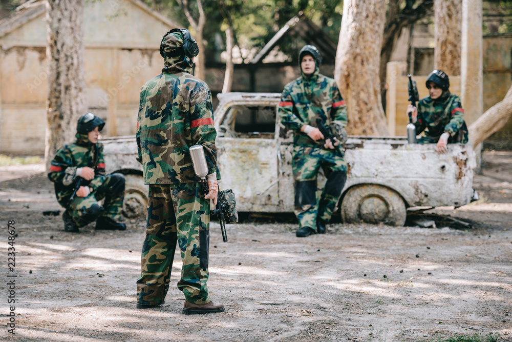Fototapeta premium paintball team in camouflage uniform with marker guns resting near broken car outdoors