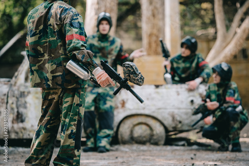 cropped image of paintball team in camouflage uniform with marker guns resting near broken car outdoors