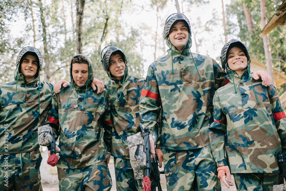 happy paintball players in camouflage with marker guns embracing each other outdoors happy paintball players in camouflage with marker guns embracing each other outdoors