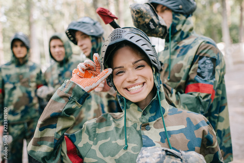 portrait of laughing female paintballer in uniform looking at camera while her team standing behind outdoors