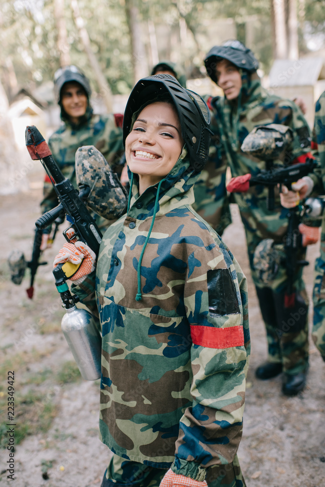 Fototapeta premium attractive smiling female paintballer in uniform holding paintball gun while her team standing behind outdoors