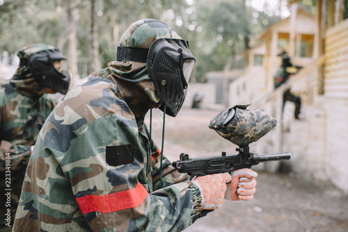 side view of male paintballer and his team in uniform and protective masks aiming by paintball guns outdoors