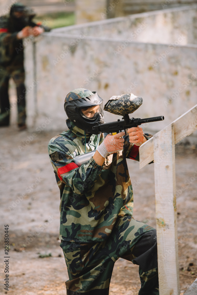 Fototapeta premium side view of confident male paintball player in goggle mask and camouflage aiming by paintball gun outdoors