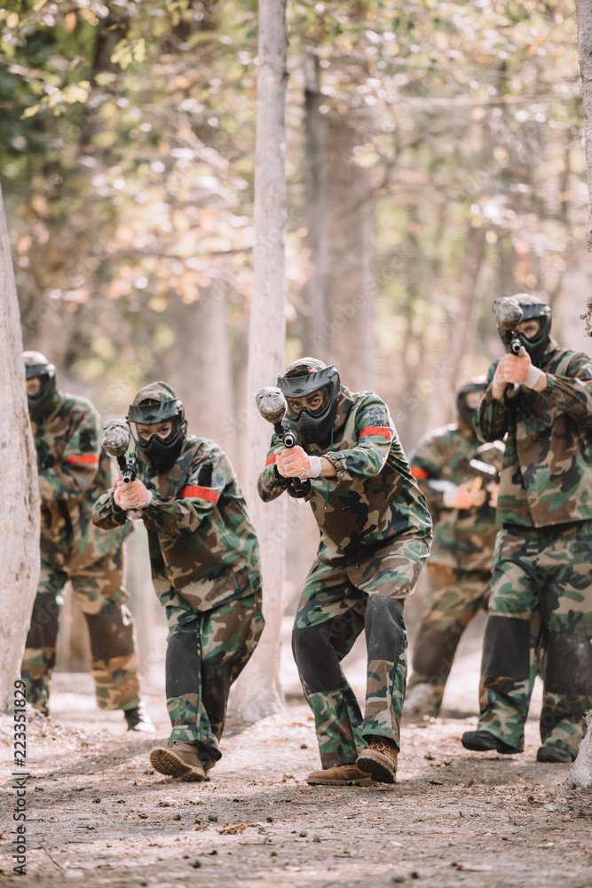 Fototapeta premium paintball team in uniform and protective masks running and aiming by paintball guns outdoors