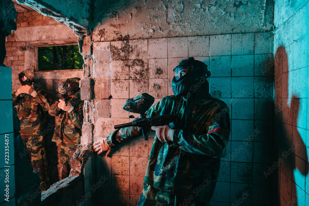 male paintball player in goggle mask and camouflage uniform hiding behind wall while other team is standing near in abandoned building