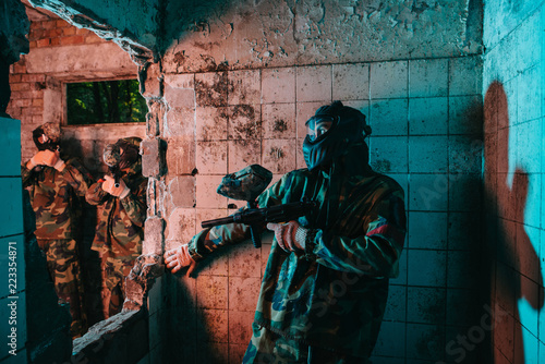 male paintball player in goggle mask and camouflage uniform hiding behind wall while other team is standing near in abandoned building © LIGHTFIELD STUDIOS