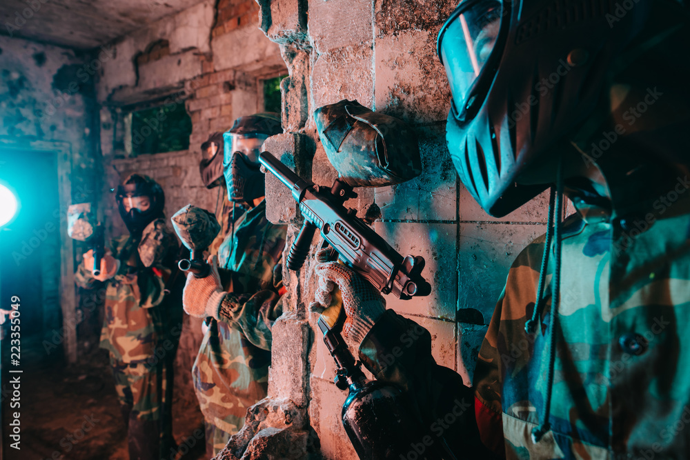 partial view of male paintball player in goggle mask and camouflage uniform hiding behind wall and aiming marker gun at player of other team in abandoned building