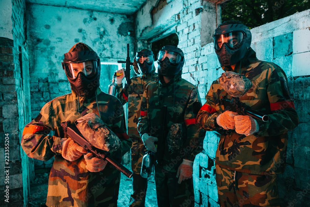 Fototapeta premium paintball team in uniform and protective masks standing with paintball guns in abandoned building
