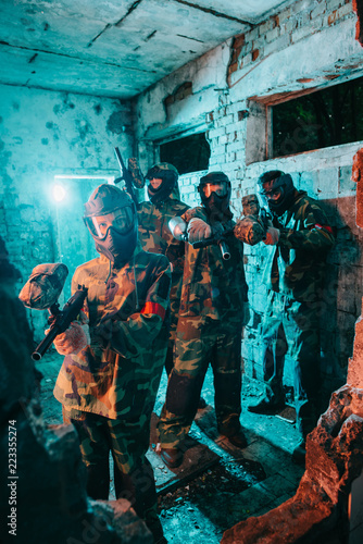 paintball team in uniform and protective masks aiming by paintball guns at camera in abandoned building