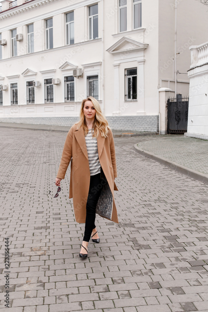 Foto Stock Beautiful young stylish blonde woman wearing long beige coat and  black high heel shoes walking through the city streets. Trendy casual outfit.  Street fashion. | Adobe Stock