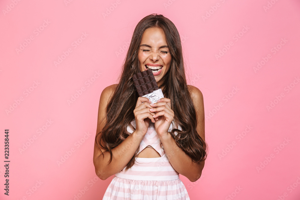 Photo of joyous woman 20s wearing dress smiling and eating chocolate bar, isolated over pink background Photo of joyous woman 20s wearing dress smiling and eating chocolate bar, isolated over pink background