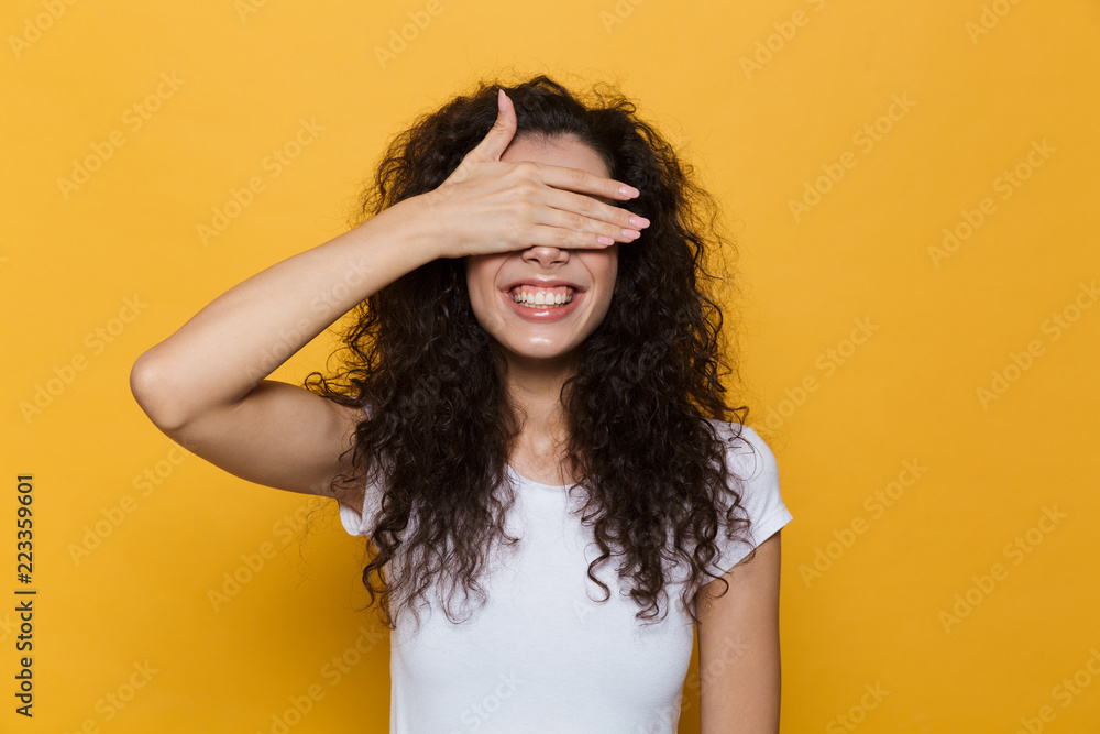 Image of brunette woman 20s with curly hair smiling and covering eyes with palm, isolated over yellow background - obrazy, fototapety, plakaty 