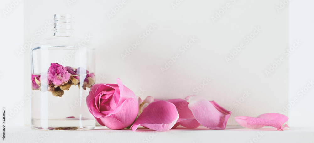 bottle of natural herbal essential oil with pink roses on white surface bottle of natural herbal essential oil with pink roses on white surface