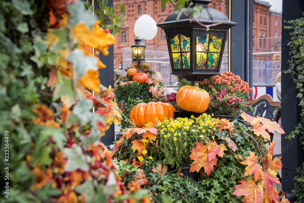 Autumn outdoor decorations. Orange pumpkin and retro forged lanterns with maple leaves,flowers and hawthorn berries in a pavillion. Red brick building