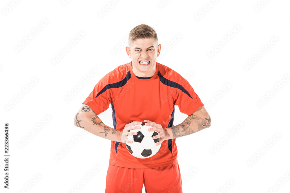 angry young soccer player holding ball and looking at camera isolated on white