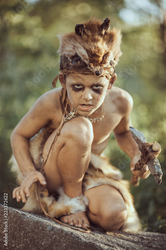 Angry caveman, manly boy with stone axe and bow hunting near river. Prehistoric tribal boy outdoors on nature. Young shaggy and dirty savage, warrior and hunter with weapon. Primitive ice age man in