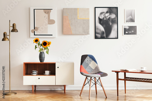 Real photo of bright eclectic living room interior with many posters, colorful chair, wooden cupboard with flowers and coffee table with book and cup photo