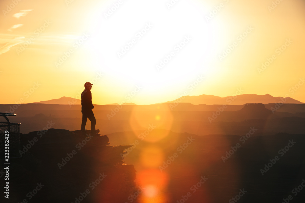 Man Silhouette On The Edge Of A Cliff Overlooking Utahs Canyonlands At Sunset With Layered Background