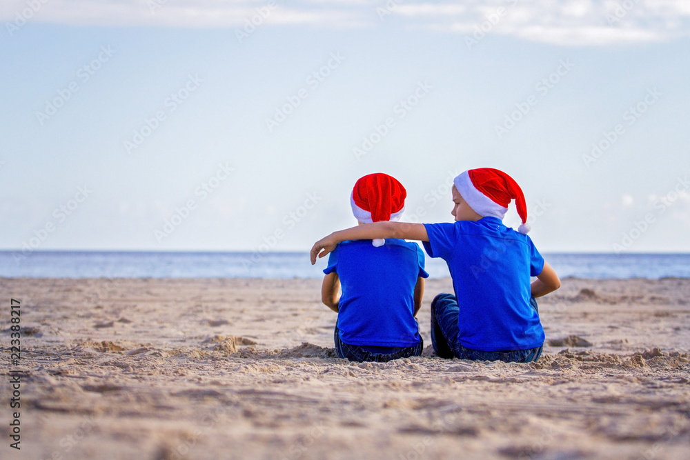 Christmas holiday. Kids in red Santa hats sit hugging at sunny beach Christmas holiday. Kids in red Santa hats sit hugging at sunny beach