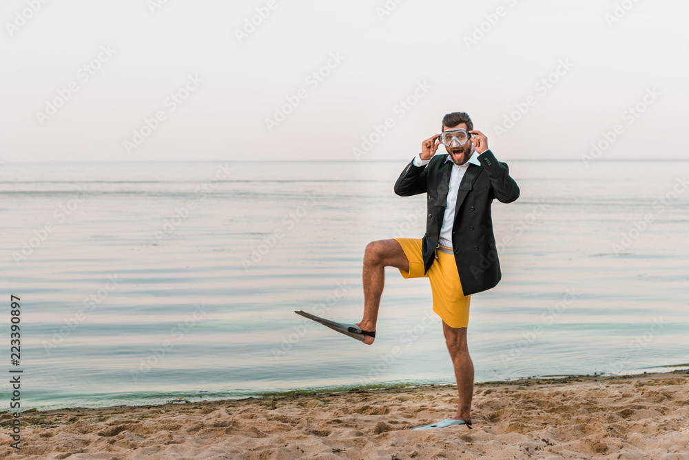 surprised man in black jacket, shorts and flippers touching swimming mask on beach near sea surprised man in black jacket, shorts and flippers touching swimming mask on beach near sea