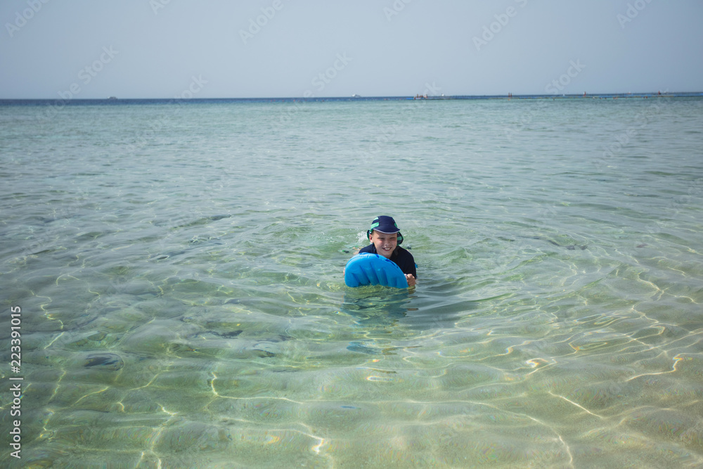 Cute white tanned kid in swimsuit, cap and aqua shoes having fun at summer sandy beach using blue inflatable surf board. Horizontal color photography.