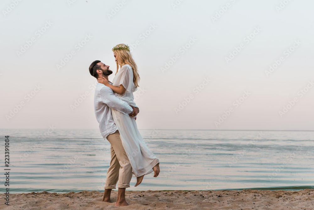 side view of groom holding bride and they looking at each other on seashore