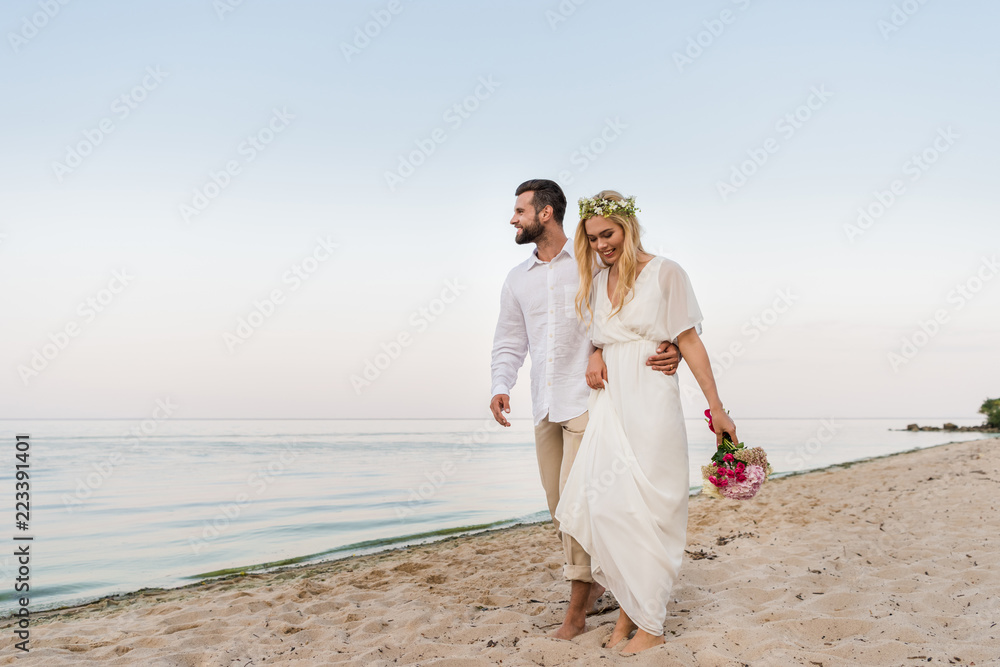 handsome groom hugging beautiful bride with wedding bouquet and they walking on seashore
