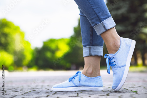 Fototapeta Naklejka Na Ścianę i Meble -  legs of a girl in jeans and blue sneakers on a sidewalk tile, a young woman strolling in a summer park