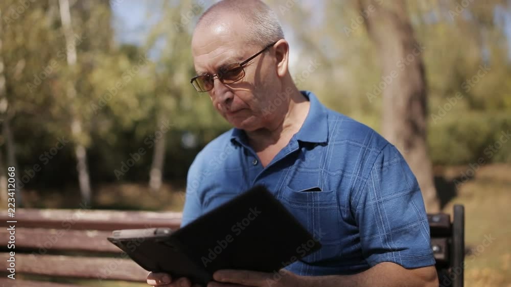 An elderly man with glasses reading an e-book on a Park bench An elderly man with glasses reading an e-book on a Park bench