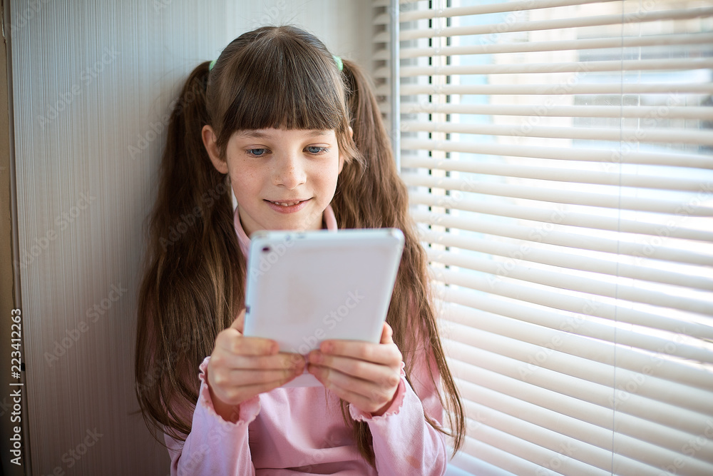 a little girl with freckles and blue eyes sitting by the window and using a tablet a little girl with freckles and blue eyes sitting by the window and using a tablet