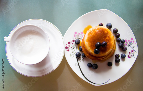 Above view of plate with cup of creamy coffee and the plate with sweet pancakes covered by blueberries fruits and maple sirup, tasteful breakfast meal close up on grey colourful background
